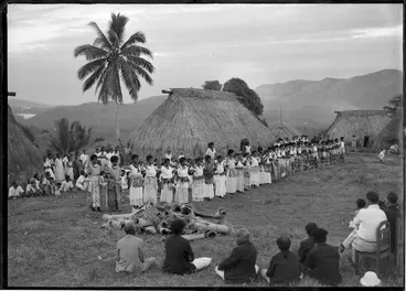 Image: Fijian women dancing, with dwellings behind, a group in the foreground watching, and another group behind the women
