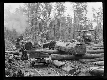 Image: Three unidentified forestry workmen loading large logs onto a steam train pulled carriages in front of a winch cable steam engine within an unknown forest location, Kakahi District, Manawatu-Whanganui Region