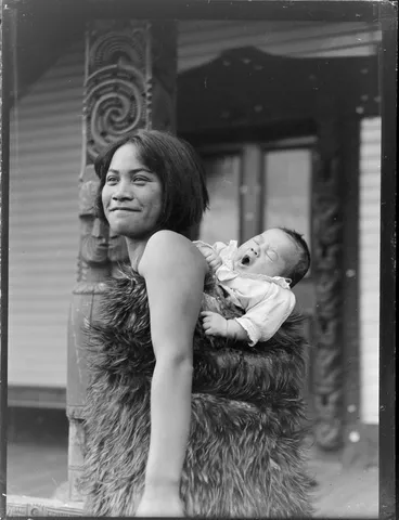 Image: Maori woman carrying a young child on her back wrapped in a feather cloak