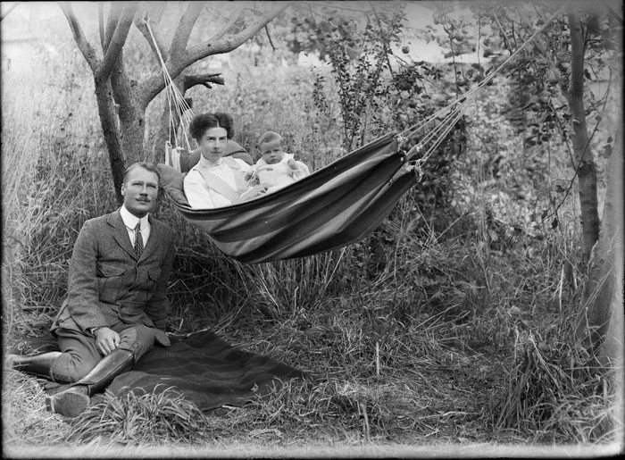 Outdoors amongst grass and trees, an unidentified family portrait of a woman and a baby with a toy trumpet in a hammock with a man with a handlebar moustache and riding attire sitting on the ground alongside, probably Christchurch region