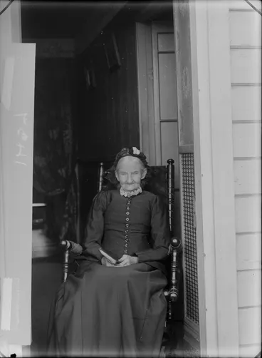 Image: Unidentified elderly woman, sitting in a rocking chair, with a book, in the doorway of a house, probably Christchurch district