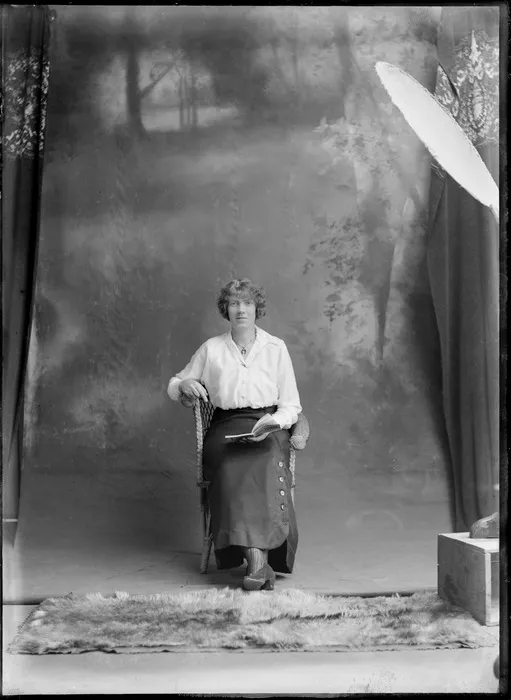 Studio portrait of unidentified woman with curly hair and horse shoe necklace sitting in cane chair reading a book, Christchurch