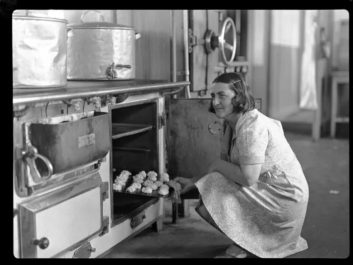Portrait of an unidentified female TEAL Catering Staff member putting a tray of scones into an oven within a kitchen, Mechanics Bay, Auckland City