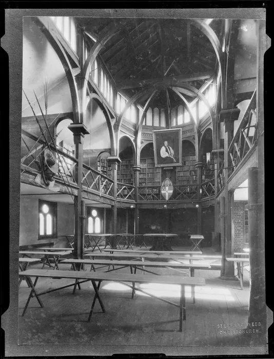 Interior of building at Christ's College, Christchurch, with shelves of books on mezanine floor, and a portrait of Bishop Henry Harper suspended from vaulted ceiling