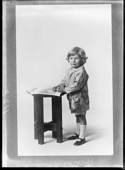 Portrait of unidentified young child standing at low table with crayon in hand and book, Christchurch