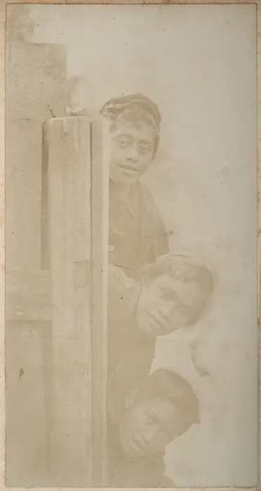 Three Maori children looking from behind a fence