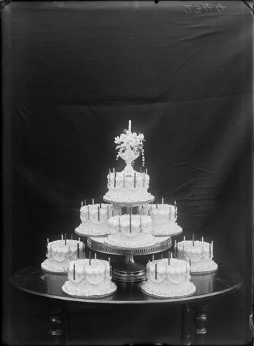 Image: Eight identical birthday cakes with candles arranged on a table, top most cake with vase holding flowers, probably Christchurch region
